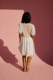 Woman wearing a beige vest over a white dress with brown boots, standing against a pink wall.