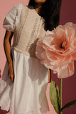 Woman wearing a white dress with lace details, standing next to a large pink flower against a pink background.