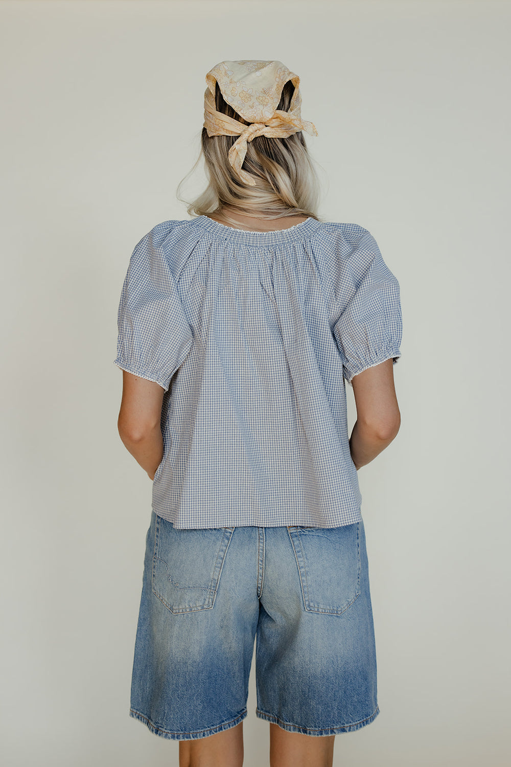 Person wearing a blue checkered top and denim shorts with a neutral headscarf on a plain background