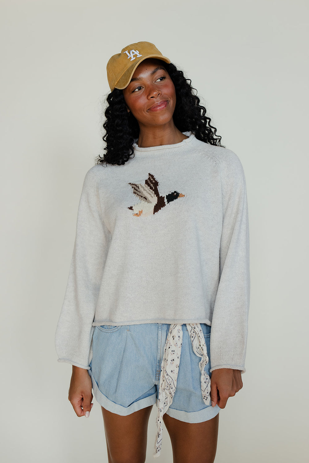 Woman wearing a white sweatshirt with a design, light blue shorts, and a yellow cap against a plain background