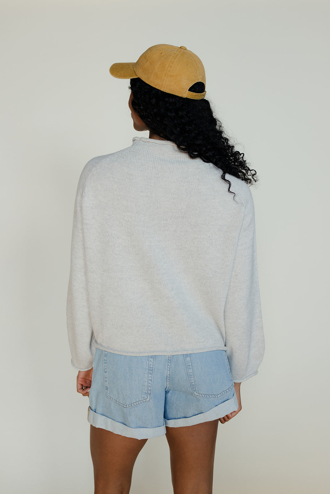 Person wearing a yellow cap, light gray sweater, and light blue shorts against a white background