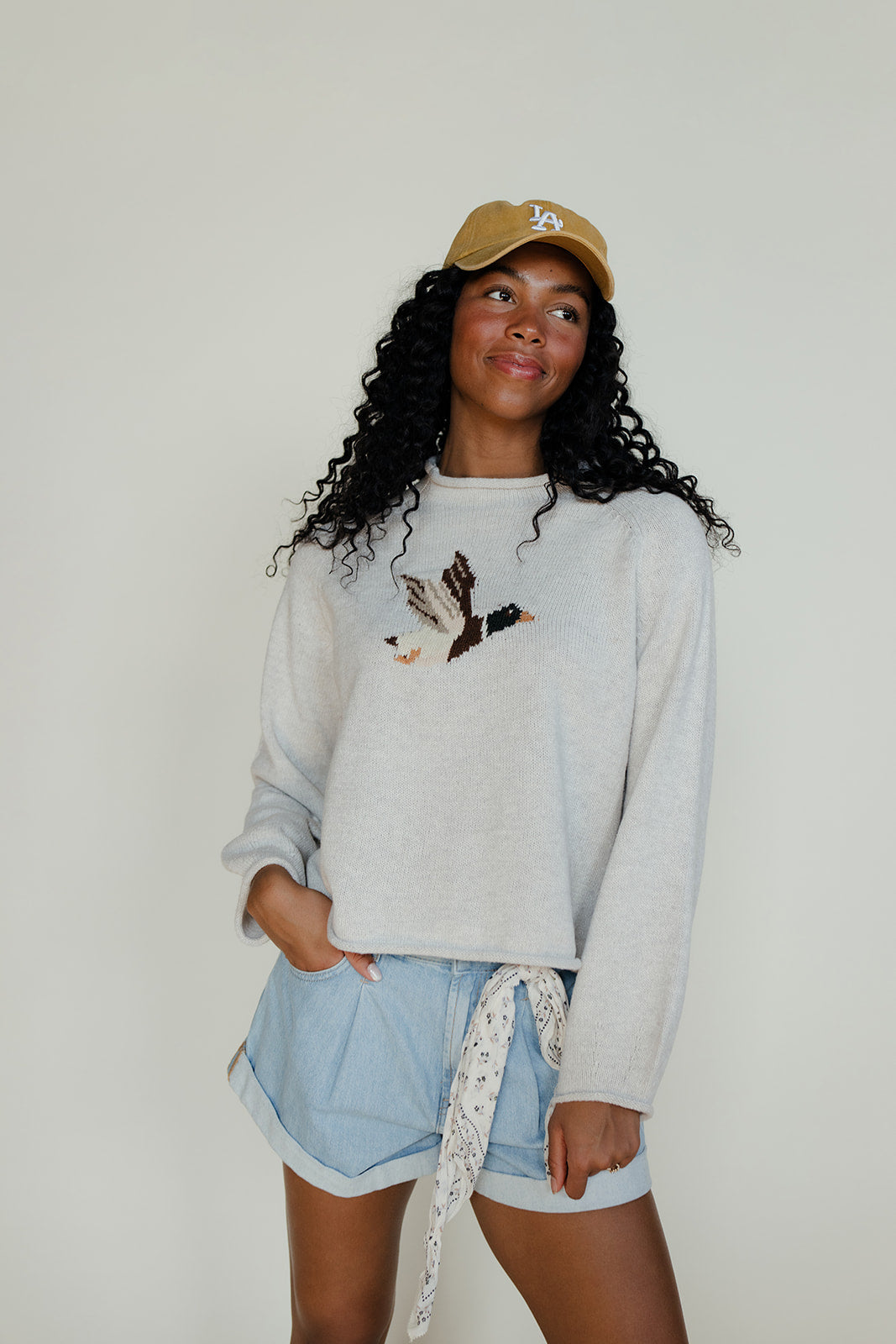 Woman wearing a light gray sweatshirt with a graphic design, light blue shorts, and a beige cap against a plain background.