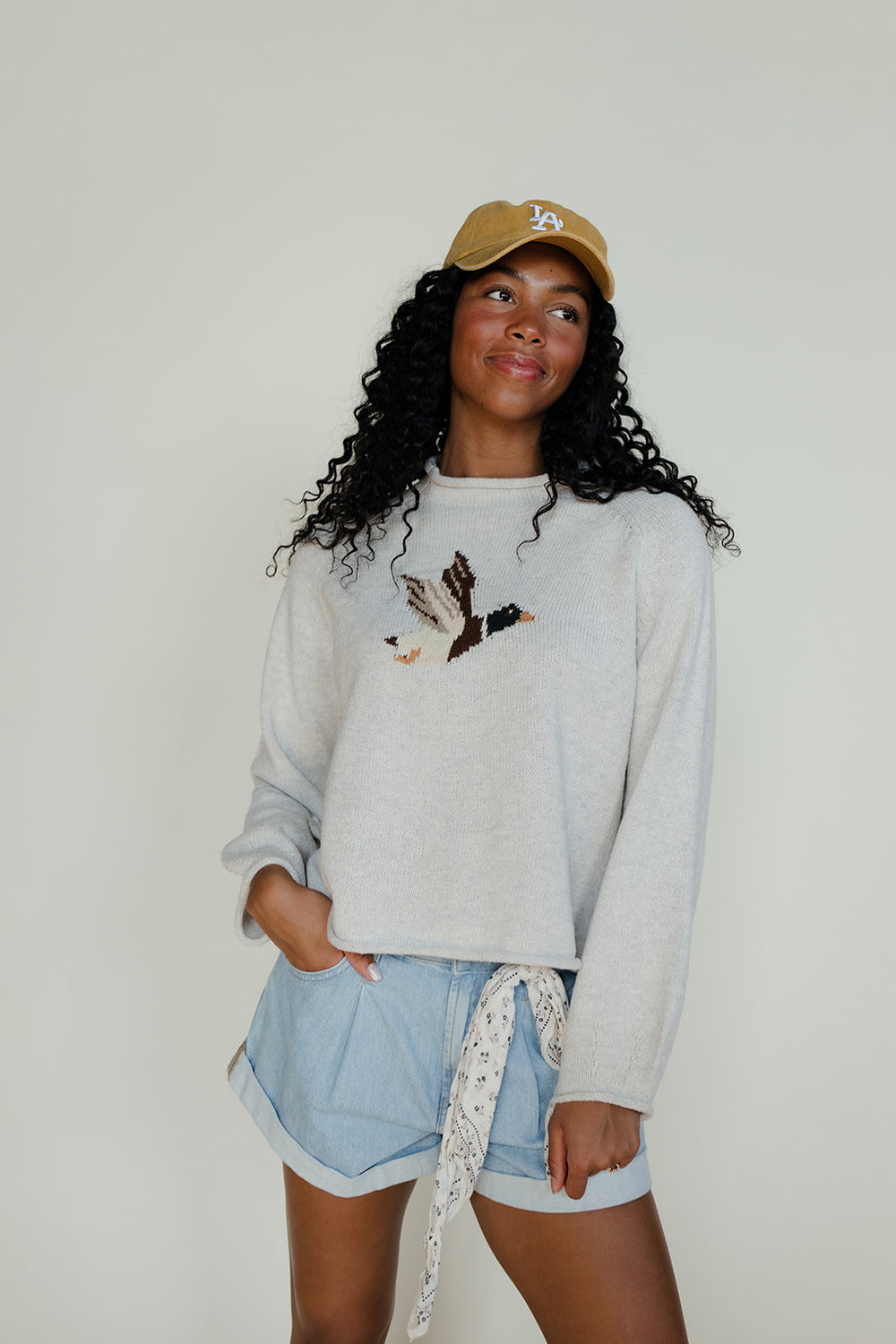 Woman wearing a light gray sweatshirt with a graphic design, light blue shorts, and a beige cap against a plain background.