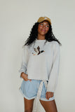 Woman wearing a light gray sweatshirt with a graphic design, light blue shorts, and a beige cap against a plain background.