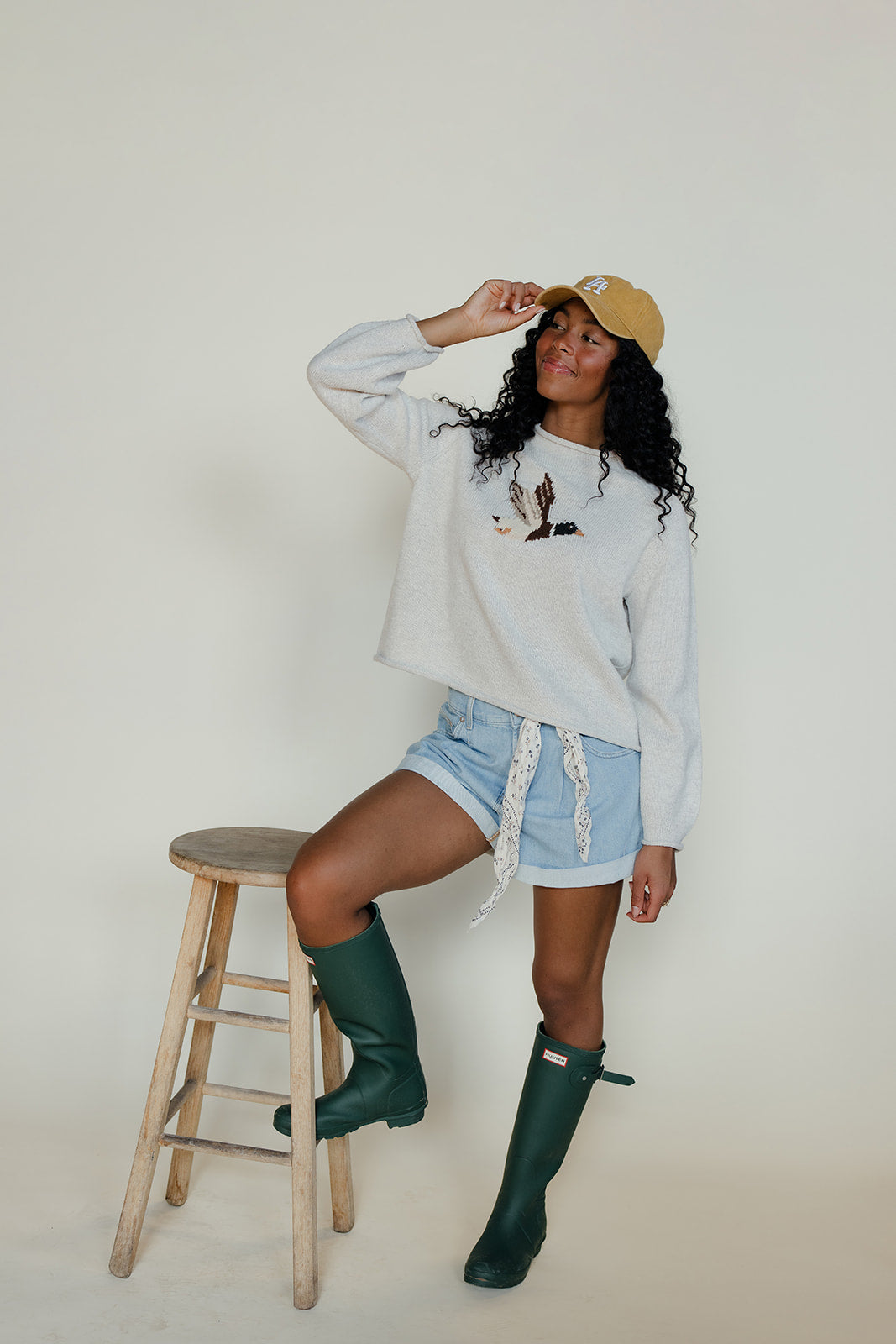Woman wearing a light gray sweatshirt, denim shorts, and green rain boots, posing on a wooden stool against a white background.