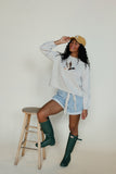 Woman wearing a light gray sweatshirt, denim shorts, and green rain boots, posing on a wooden stool against a white background.