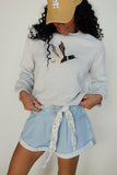 Person wearing a light blue sweater with a design, denim shorts, and a beige cap with 'LA' logo.