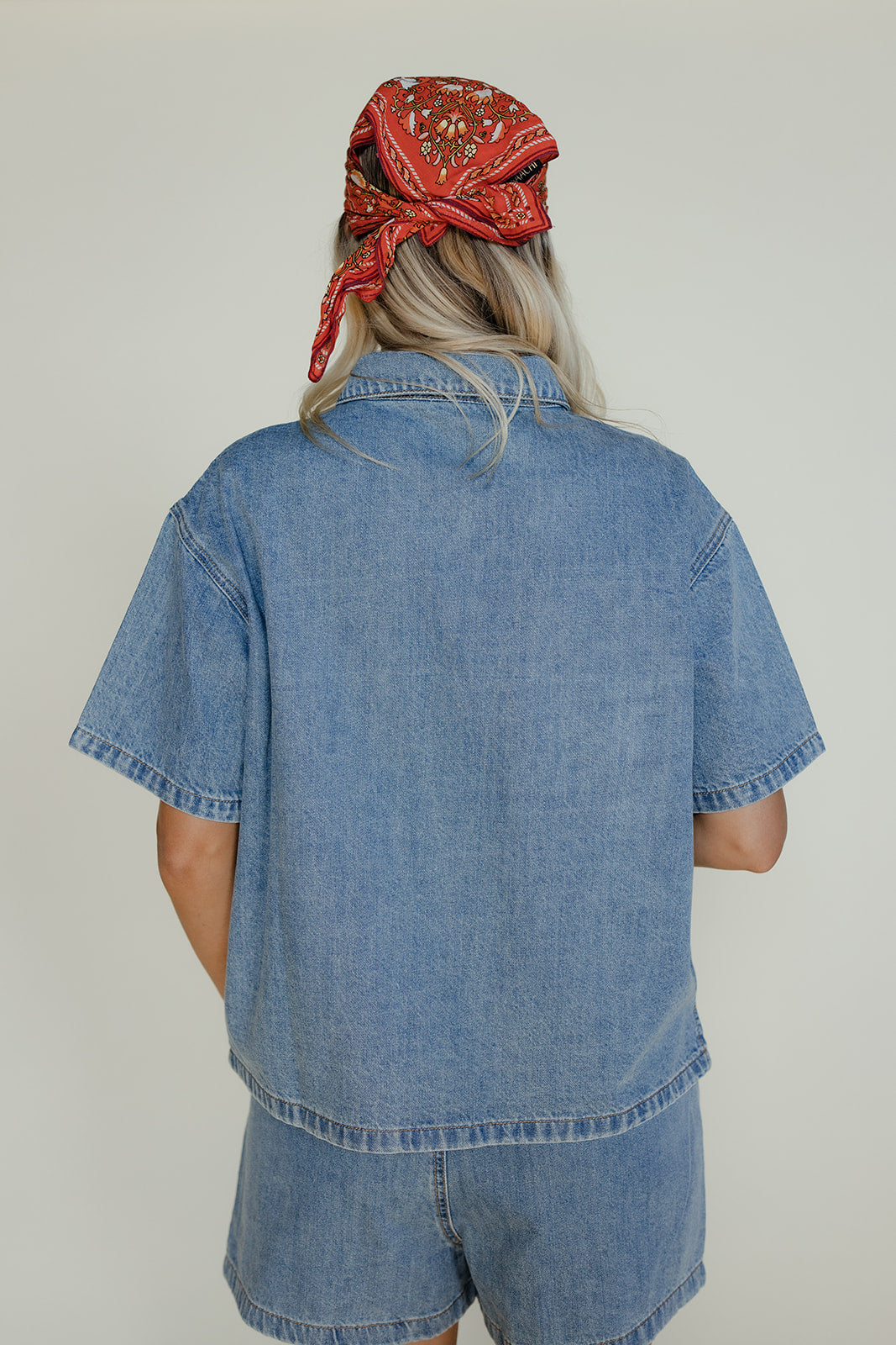 Person wearing a blue denim shirt and shorts with a red bandana on a plain background