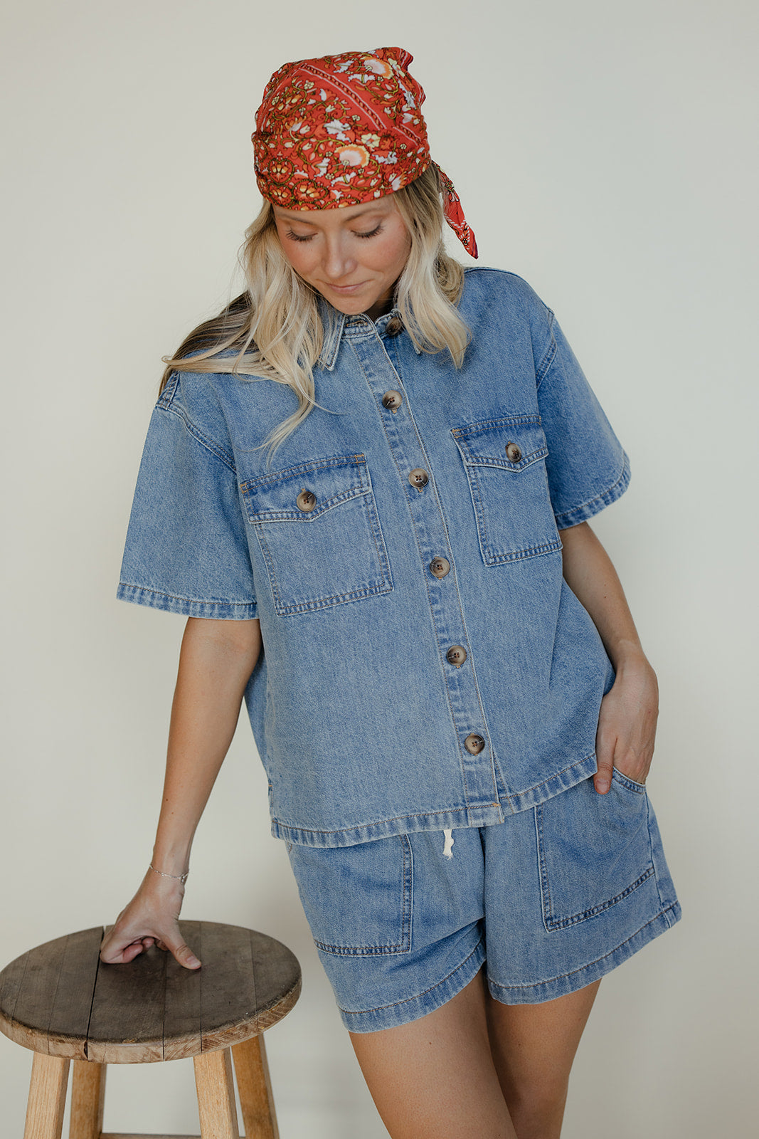 Woman wearing a denim outfit with a red patterned headscarf, standing next to a wooden stool.