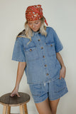 Woman wearing a denim outfit with a red patterned headscarf, standing next to a wooden stool.
