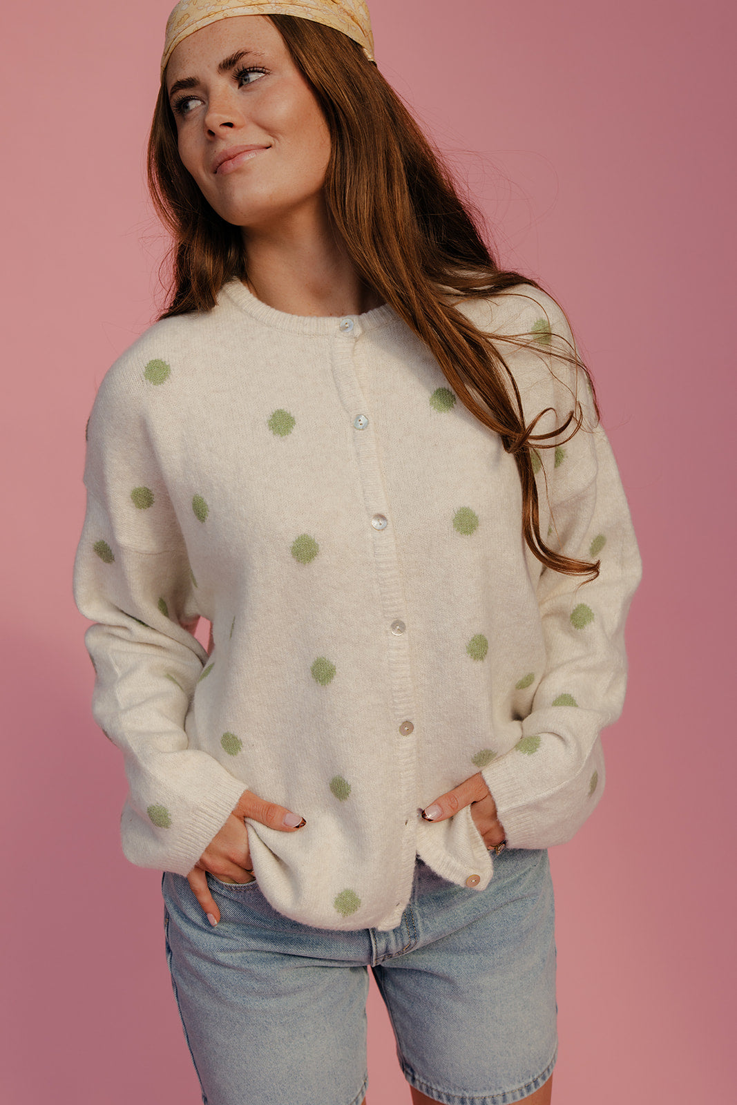 Woman wearing a white cardigan with green polka dots and denim shorts on a pink background
