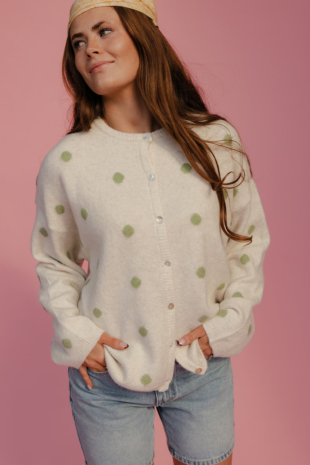 Woman wearing a white cardigan with green polka dots and denim shorts on a pink background