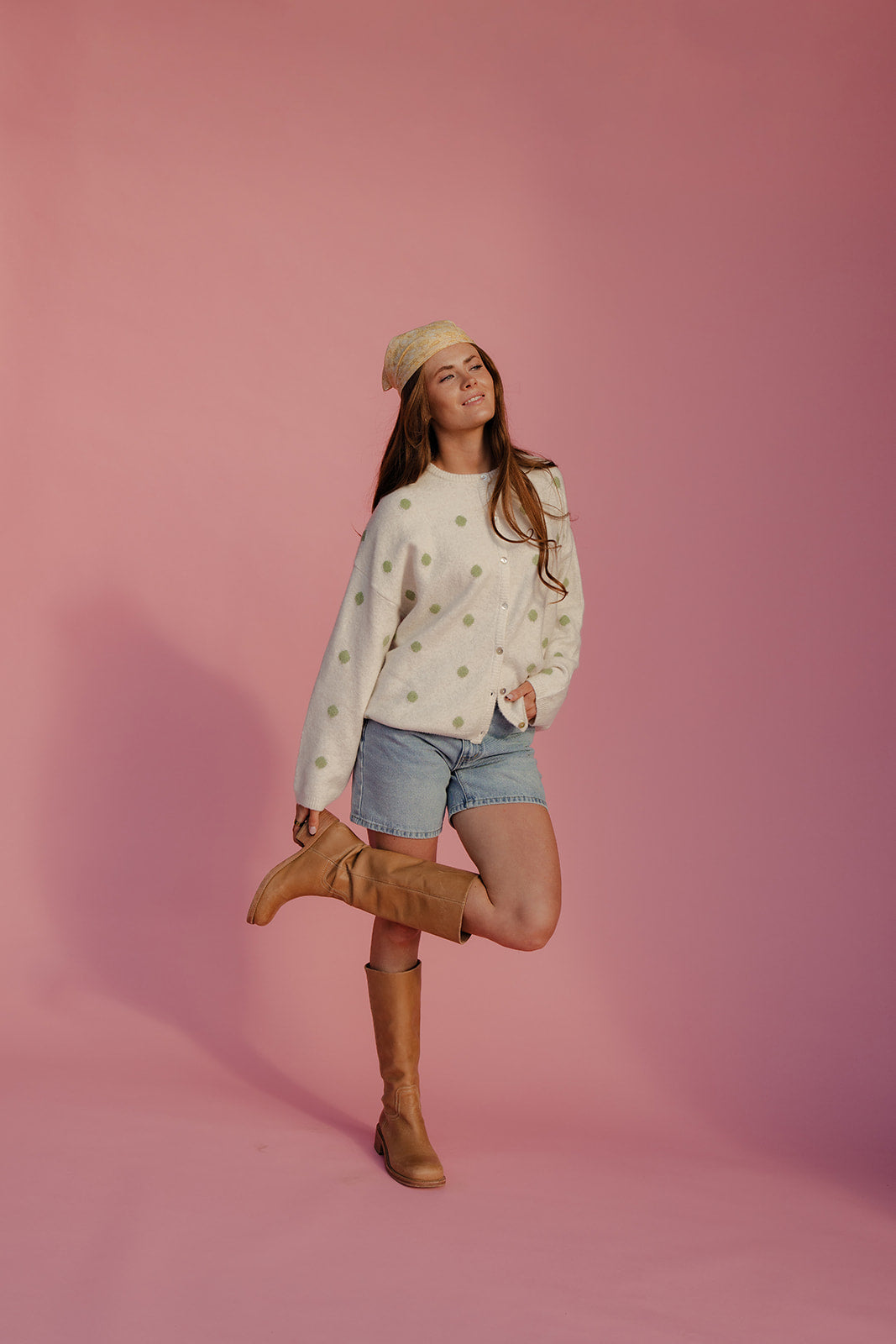 Woman posing in a polka dot sweater, denim shorts, and brown boots against a pink background