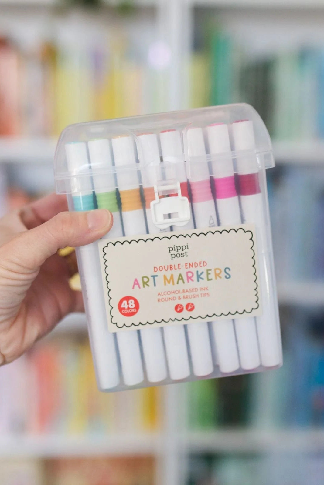 Pack of double-ended art markers held by a hand with a blurred bookshelf background
