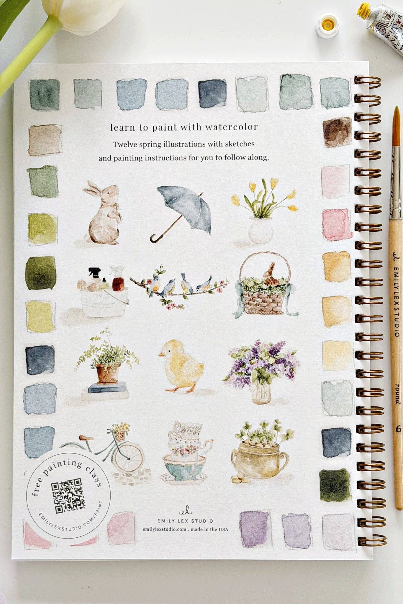 Art book with watercolor illustrations on a white surface with a plant and paint bottle.