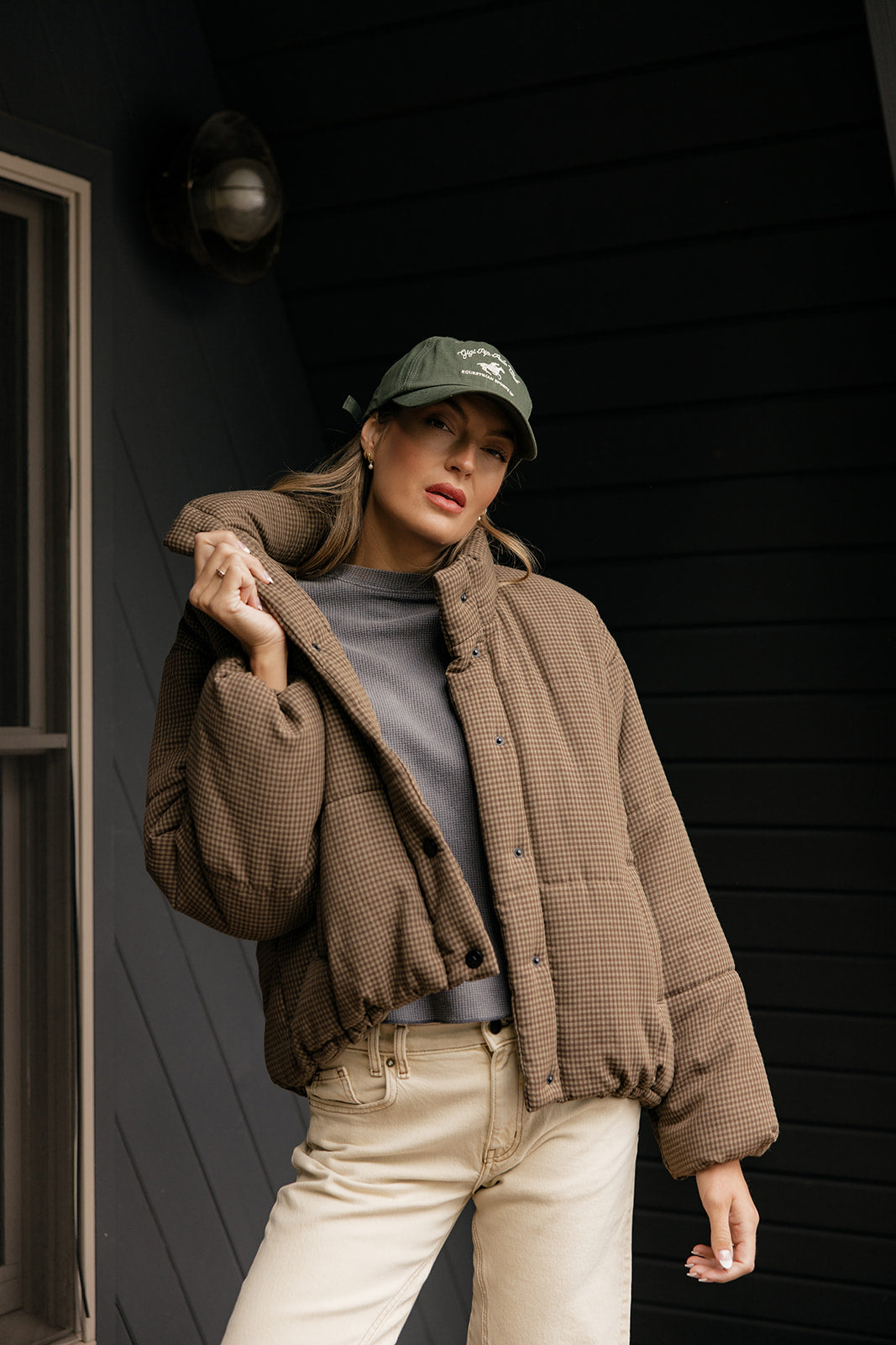 Person wearing a brown jacket, gray shirt, beige pants, and green cap against a dark background.