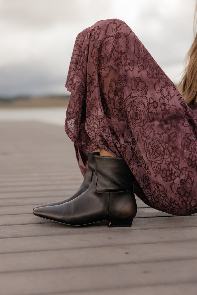 Person wearing black boots and a patterned skirt sitting on a wooden deck.