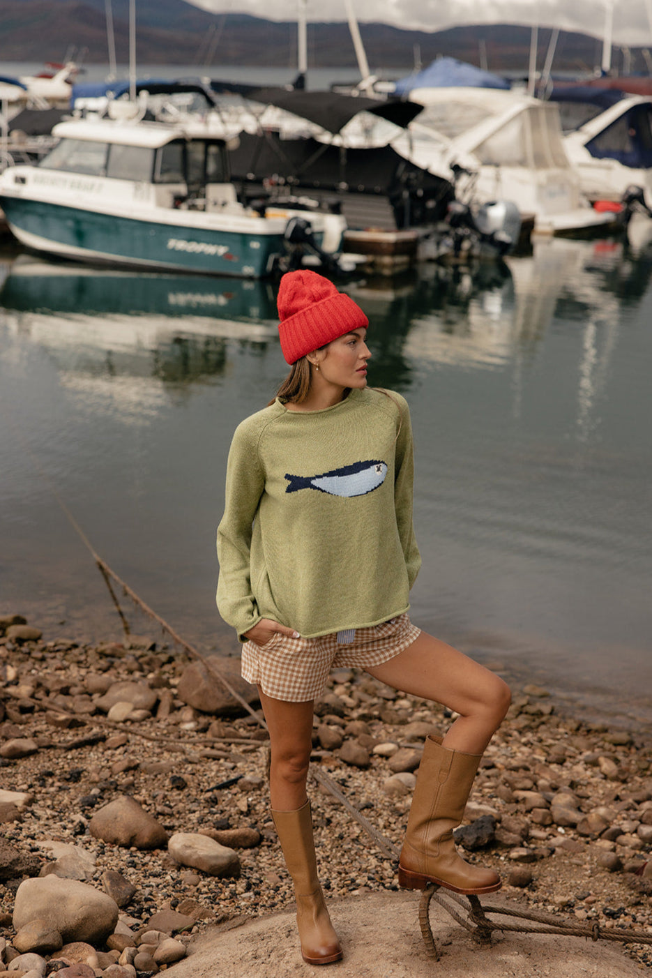 Person wearing a green sweater with a fish design, red beanie, and plaid shorts standing on rocks by a marina.