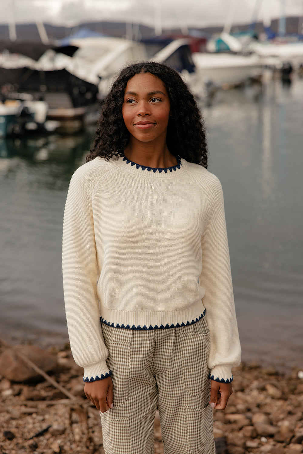 Woman standing by a marina wearing a cream sweater with navy trim and checkered pants.