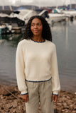 Woman standing by a marina wearing a cream sweater with navy trim and checkered pants.