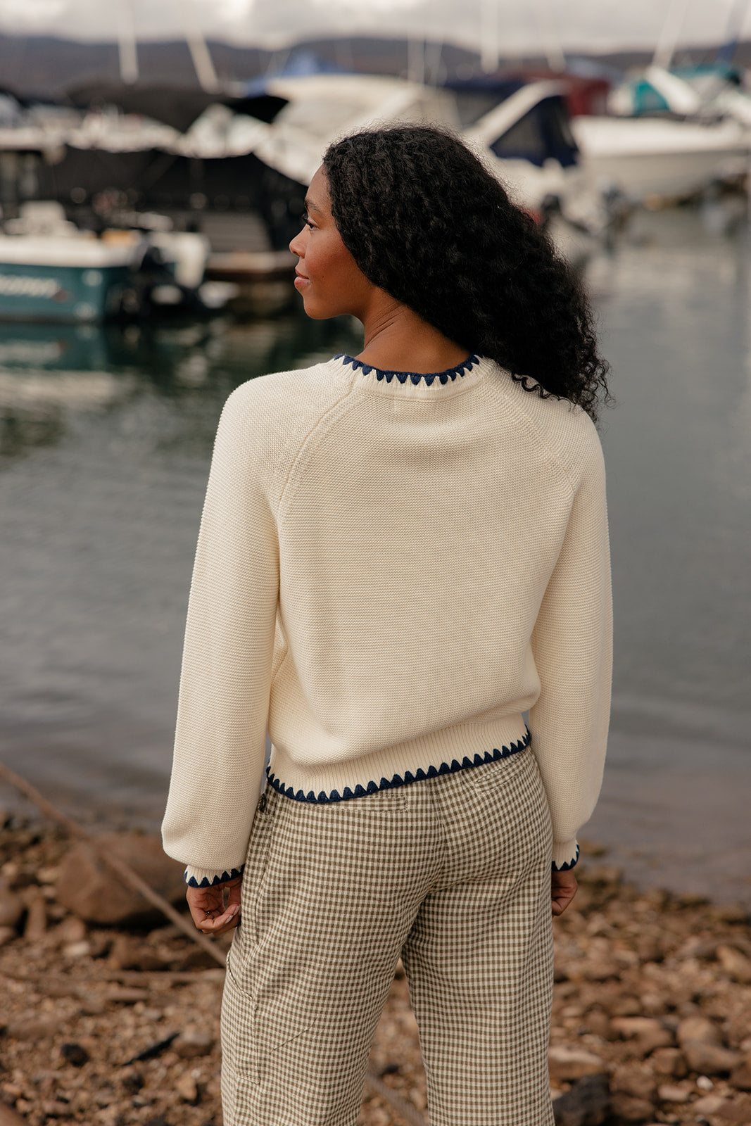 Woman standing by a marina wearing a cream sweater and checkered pants.