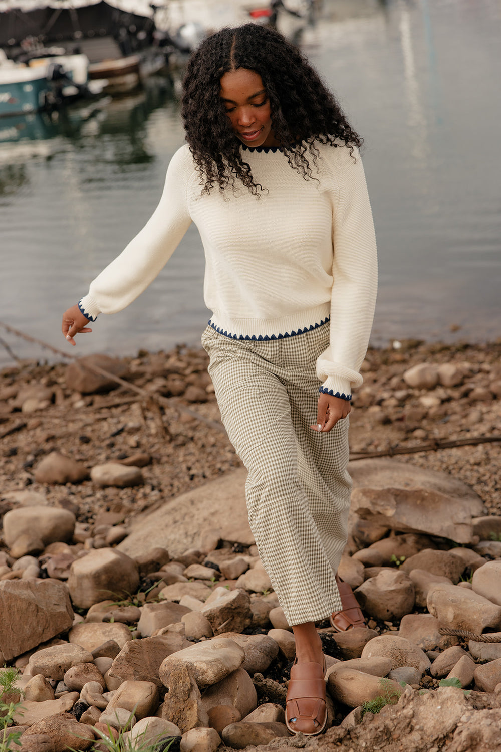 Woman walking on rocks by a body of water wearing a white sweater and patterned pants.