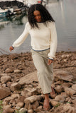 Woman walking on rocks by a body of water wearing a white sweater and patterned pants.