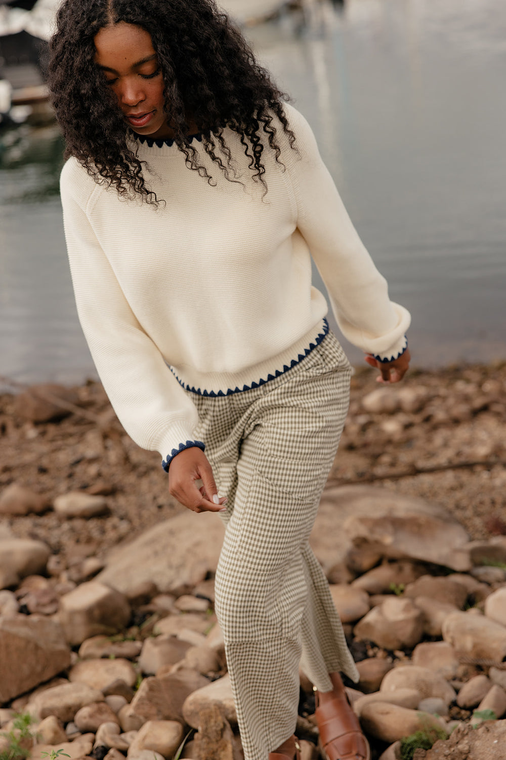 Woman in a cream sweater and patterned skirt standing on rocky terrain by a body of water.