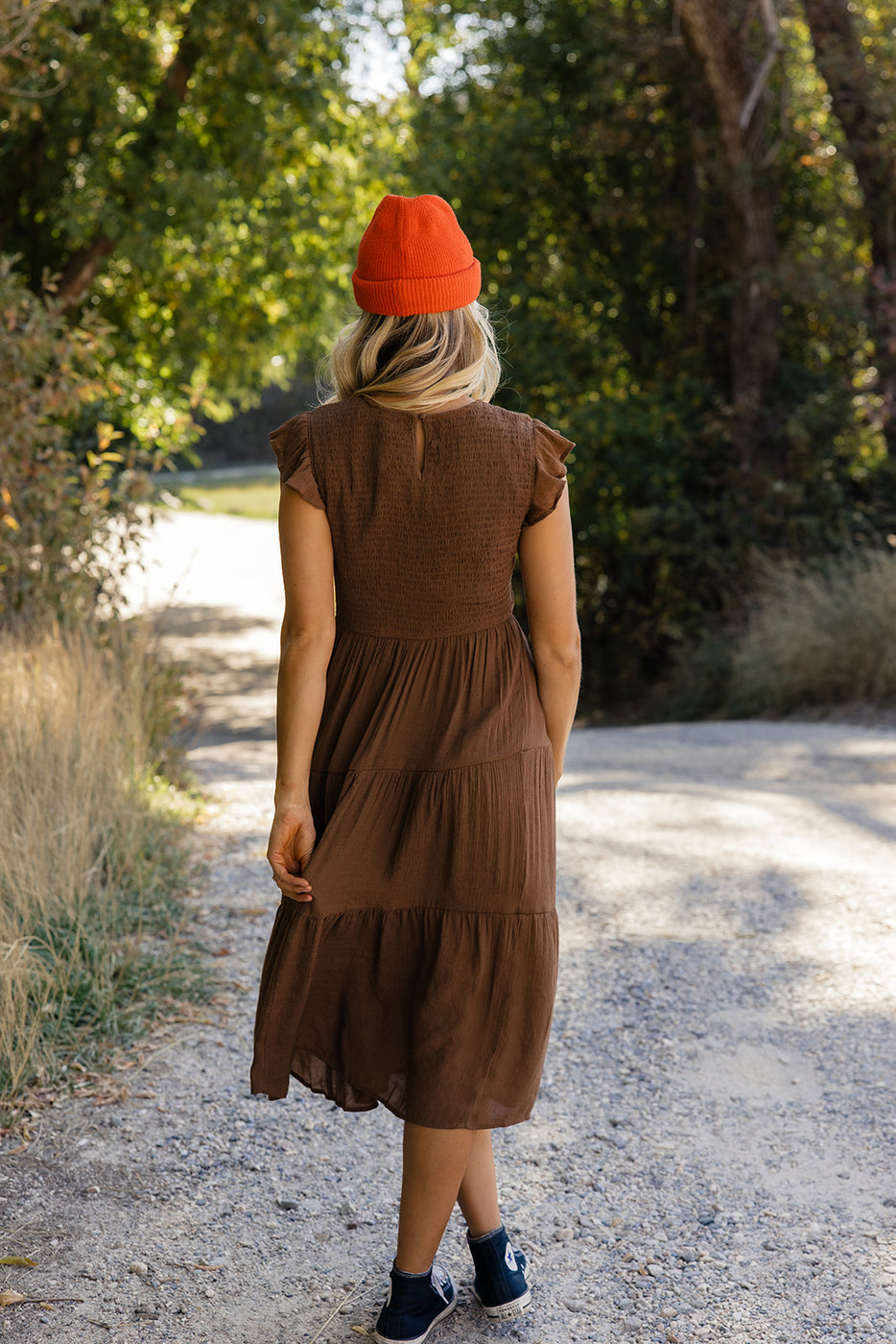 a woman wearing a brown dress and orange hat walking on a gravel path