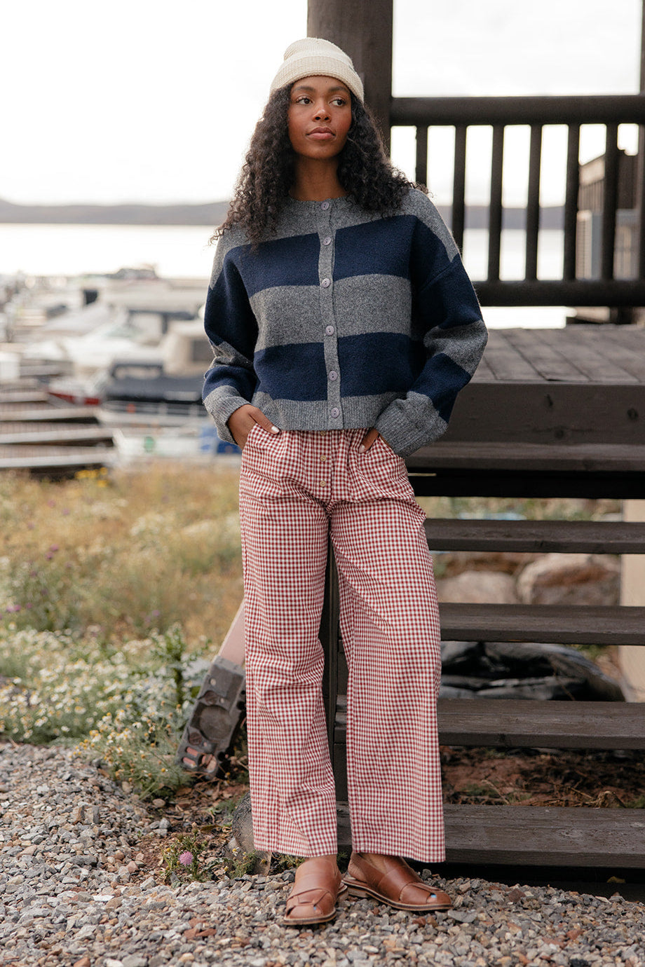 Woman wearing a patterned sweater and pink checkered pants standing on a wooden deck with a scenic background.
