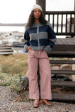 Woman wearing a patterned sweater and pink checkered pants standing on a wooden deck with a scenic background.
