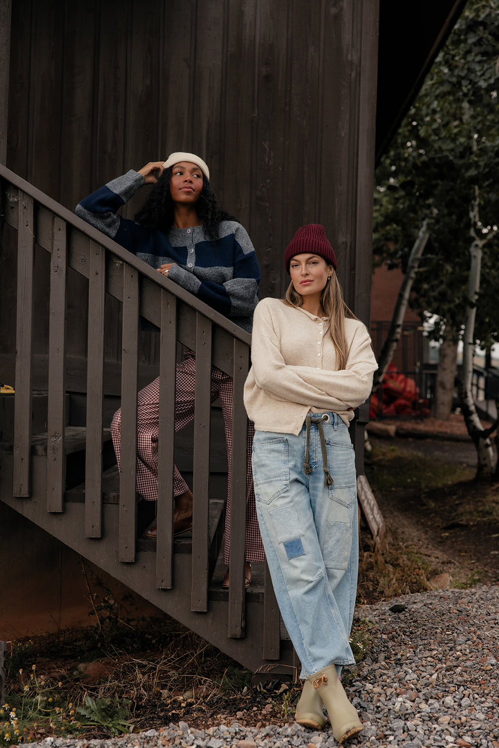 Two women in a rustic outdoor setting, one standing on steps and the other standing on the ground.