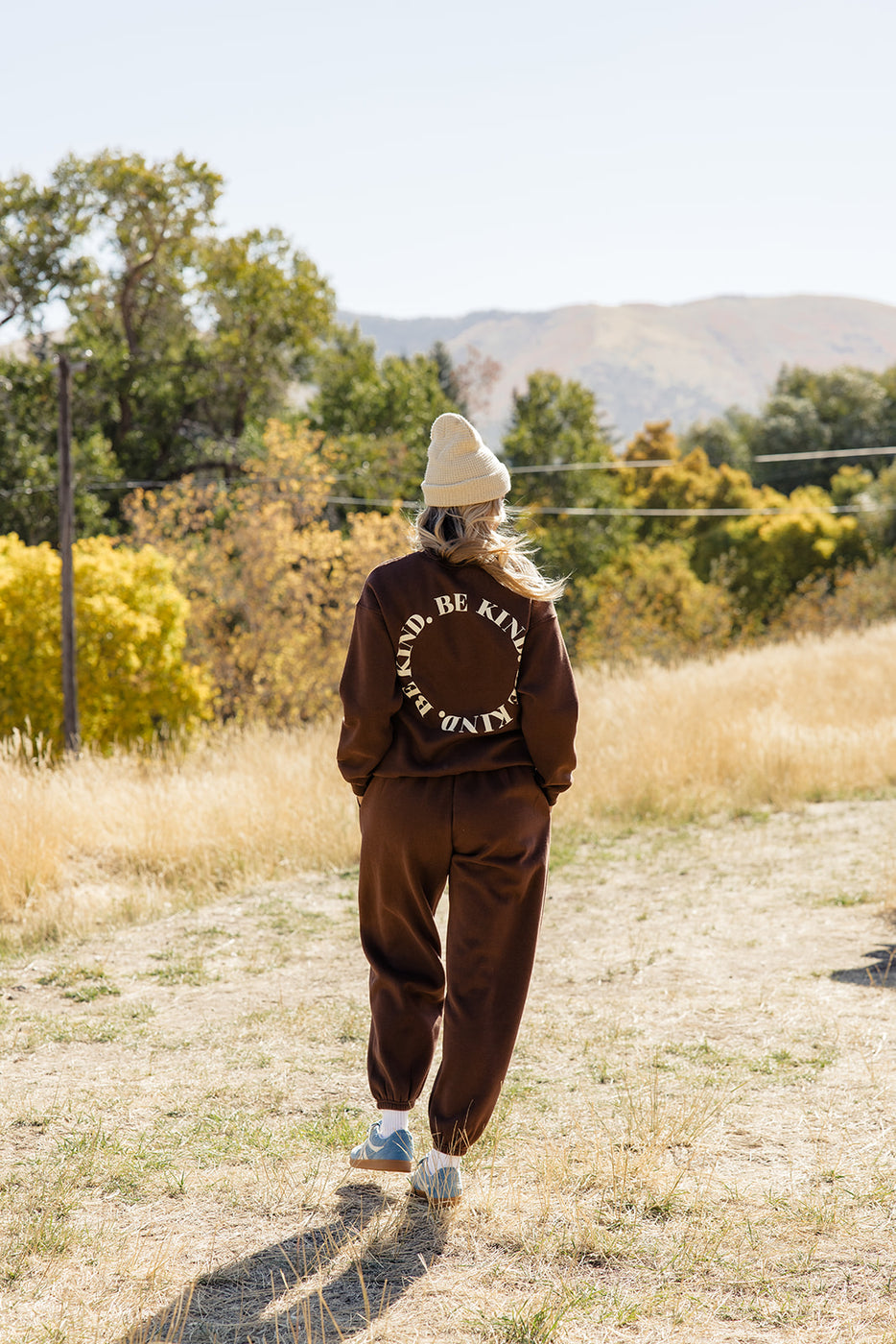 a woman wearing a brown sweater and hat walking on a dirt path