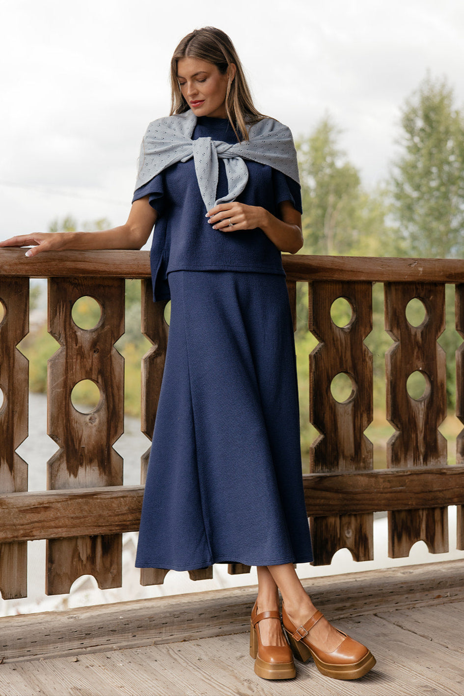 Woman in a blue dress with a gray shawl standing on a wooden deck.