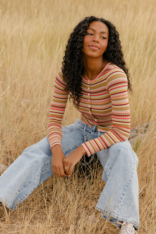 Woman sitting in a field of tall grass wearing a striped sweater and jeans.