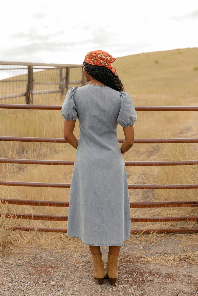 Woman in a light blue dress and brown boots standing near a metal fence with a field in the background.