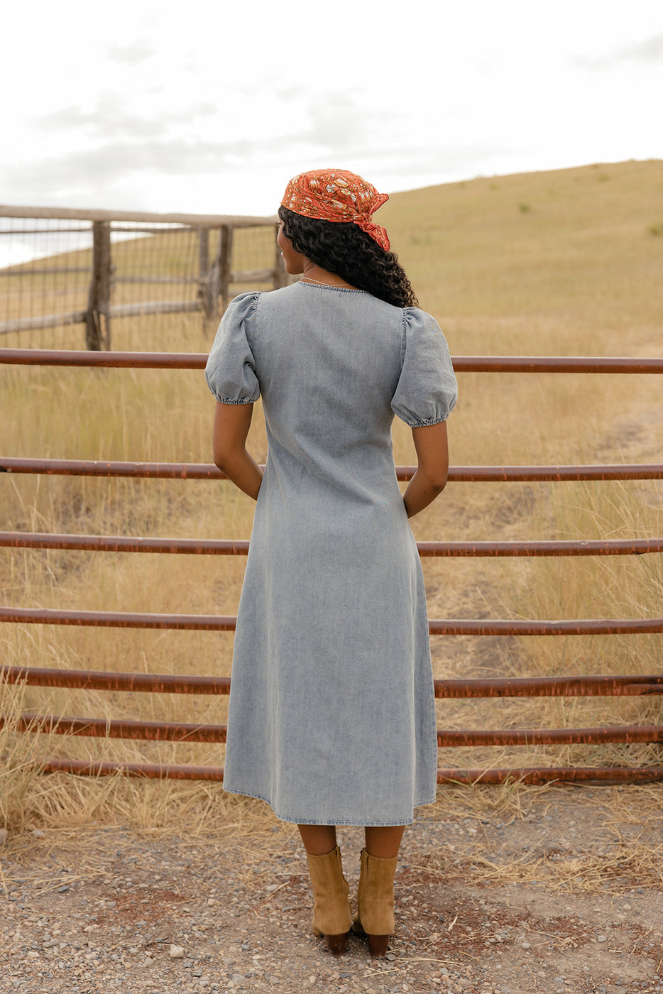 Woman in a light blue dress and brown boots standing near a metal fence with a field in the background.