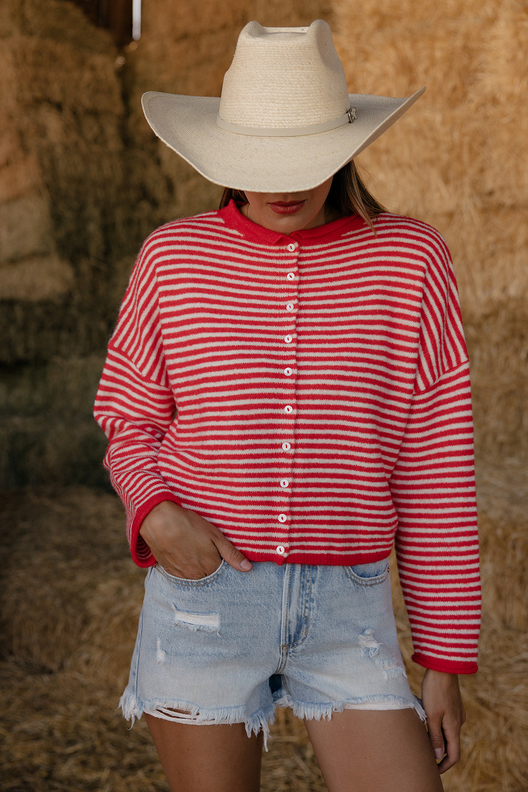 Person wearing a red and white striped shirt, denim shorts, and a cowboy hat in front of hay bales.