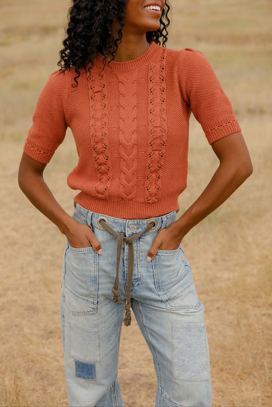 Woman wearing a rust-colored knitted sweater and light blue jeans against a blurred natural background