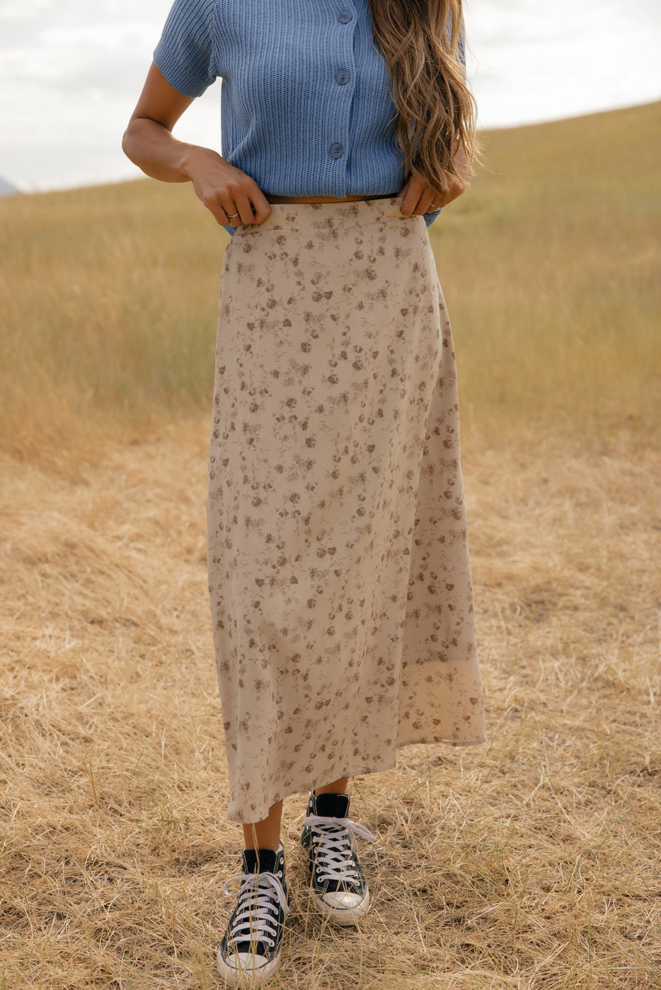 Person wearing a blue top and beige floral skirt standing in a field.