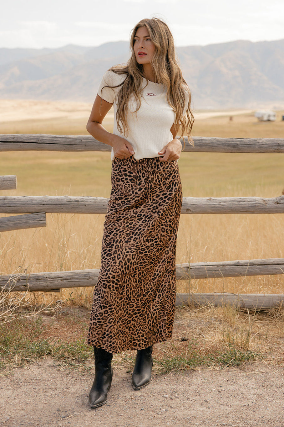 Woman in a white top and leopard print skirt standing in a field with mountains in the background