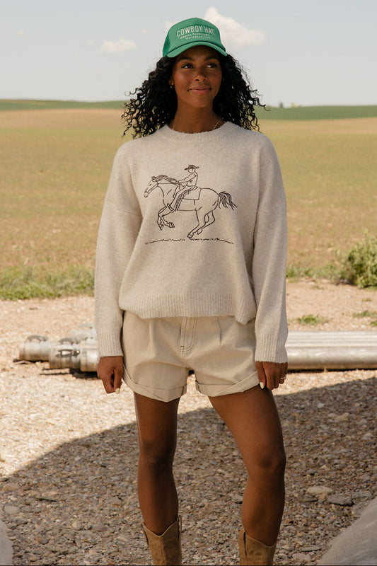Woman wearing a sweater with a horse graphic and a green cap in an open field.