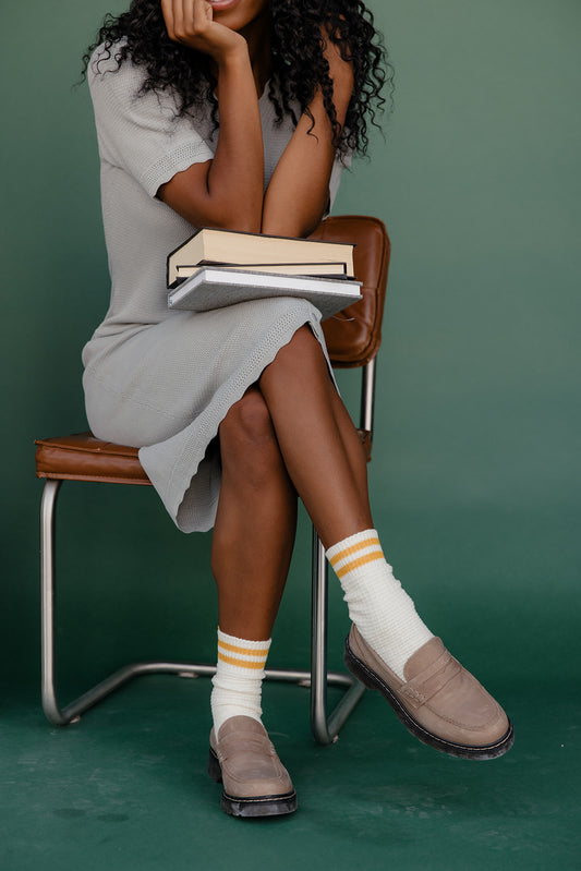 Woman sitting on a chair holding books, wearing white socks with yellow stripes and brown shoes.