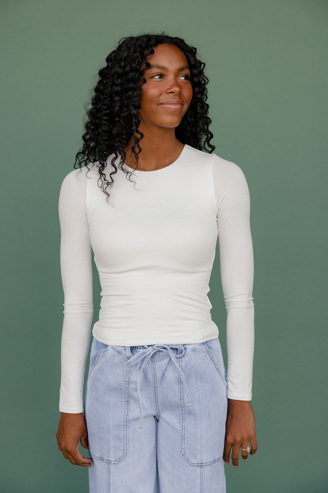 Woman wearing a white long-sleeve top and light blue jeans against a green background