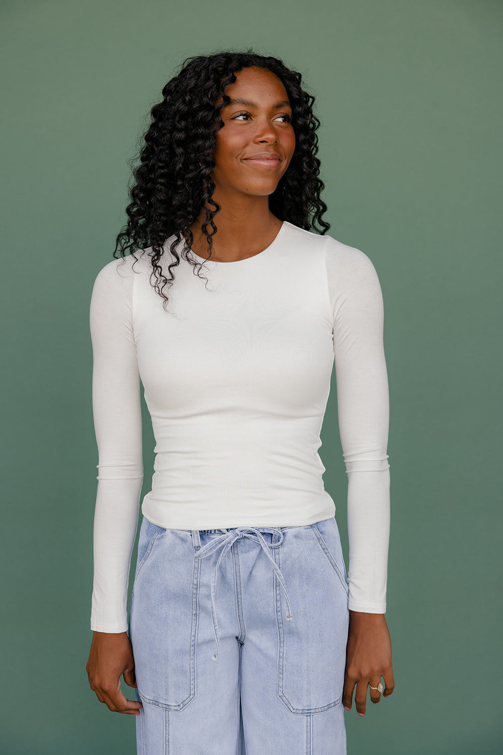 Woman wearing a white long-sleeve top and light blue jeans against a green background
