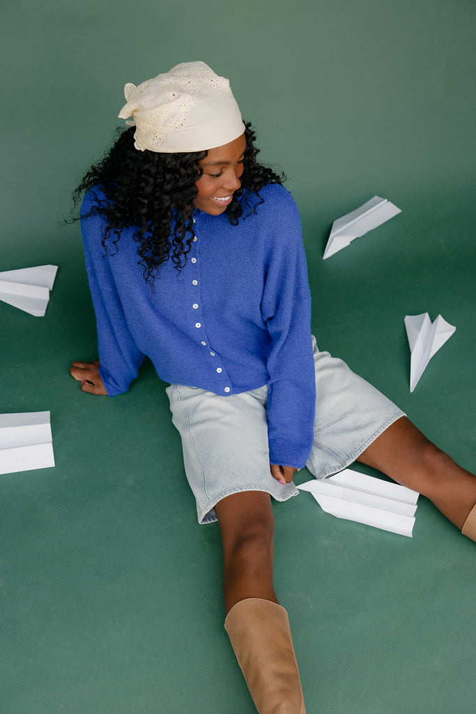 Woman sitting on a green floor with paper airplanes around her, wearing a blue shirt and white shorts.