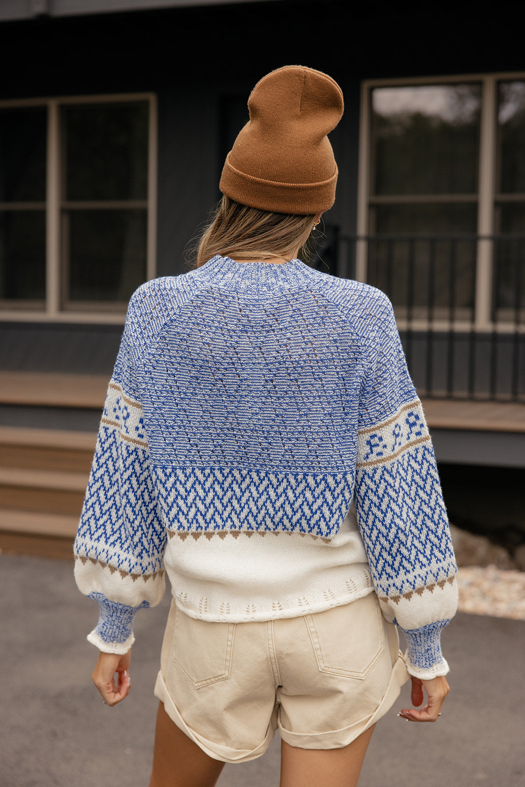 Person wearing a blue patterned sweater and beige shorts, standing in front of a building.