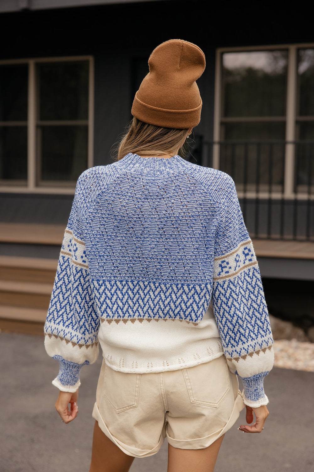 Person wearing a blue patterned sweater and beige shorts, standing in front of a building.
