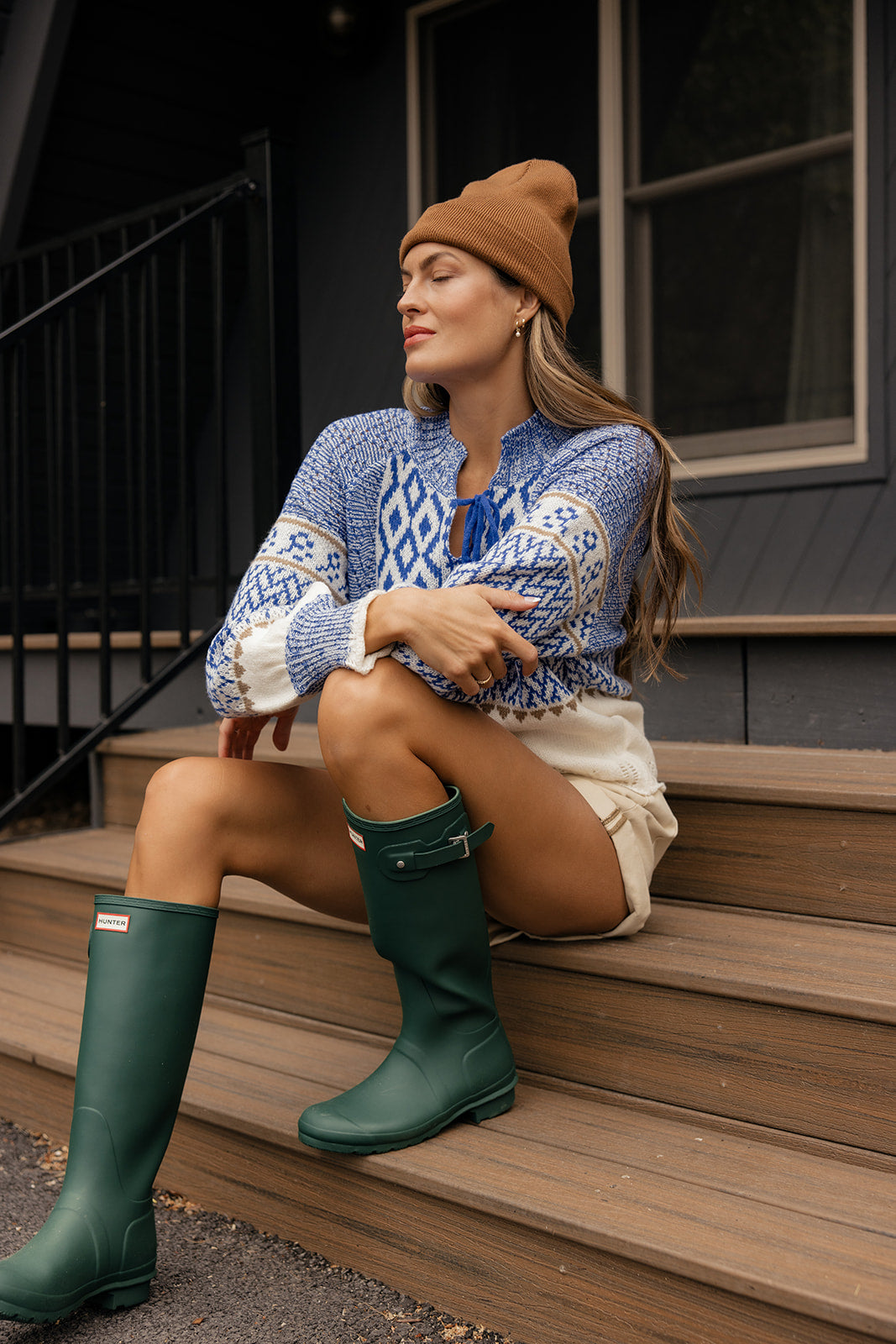 Woman sitting on steps wearing a blue patterned sweater, beige skirt, brown beanie, and green rain boots.