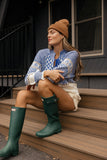 Woman sitting on steps wearing a blue patterned sweater, beige skirt, brown beanie, and green rain boots.